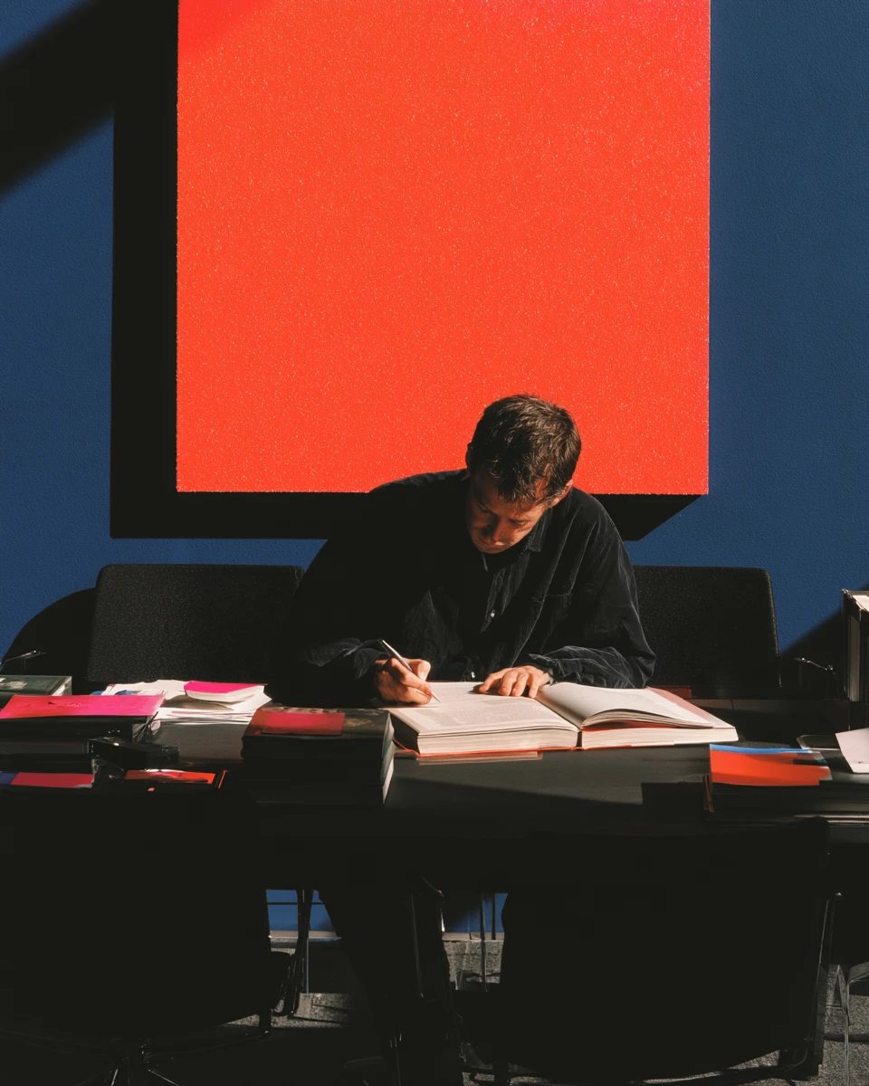 A man writing at a desk of open books beneath a large red canvas